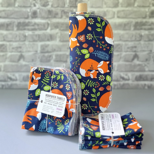 Navy Foxes print NonPaper Towels. The print is bright orange foxes with their cubs, various leaves, flowers, small branches, and acorns in autumnal colors on the navy blue background. In the center, they are rolled onto a wooden paper towel holder, and there are bundled packs of 10 and 5 sitting next to the holder. The bundles are tied with string and have a small paper tag. The background is a gray brick wall and solid gray tabletop. 