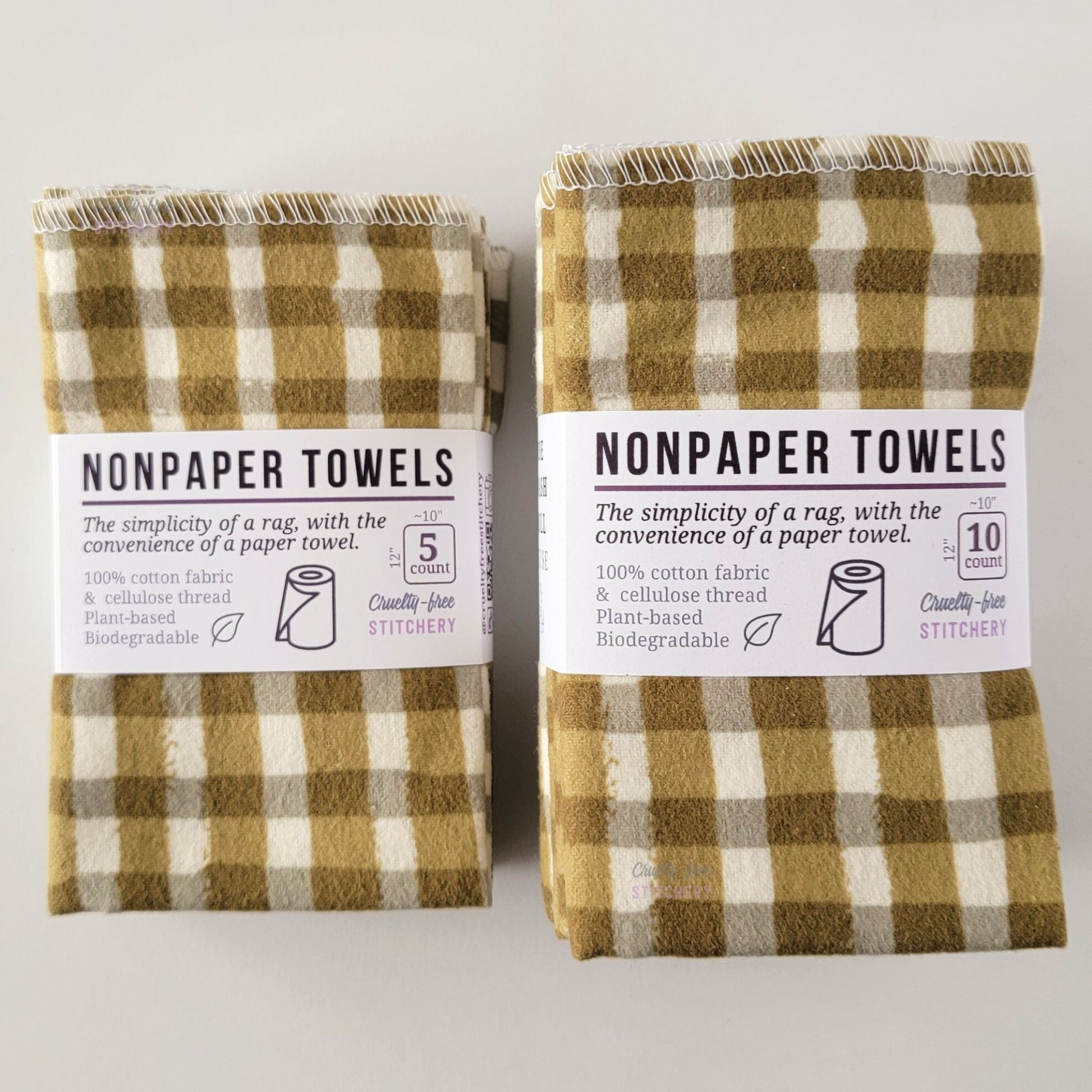 Two packages of the Olive Painted Plaid NonPaper Towels, one 5-pack and one 10-pack. Both wrapped with a paper tag, The tag also has the fiber content, a graphic of a paper towel roll, the Cruelty-Free Stitchery logo, and a rectangle with rounded corners that has the dimensions on the outside, 10x12", and the number of towels inside.