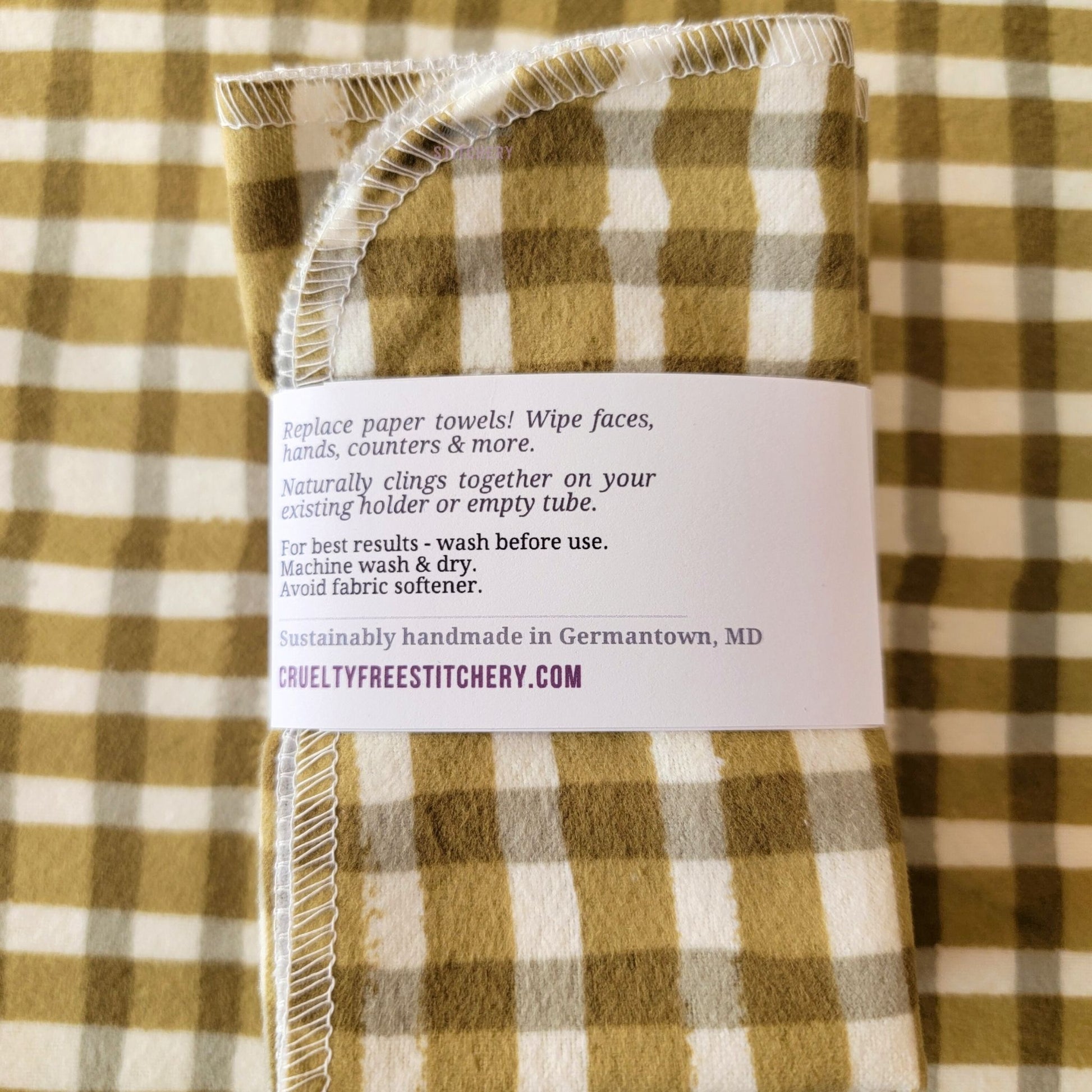 The back of a packaged bundle of the Olive Painted Plaid NonPaper Towels. The tag reads "Replace paper towels! Wipe faces, hands, counters, and more. Naturally clings together on your existing holder or empty tube. For best results - wash before use. Machine wash and dry. Avoid fabric softener. Sustainable handmade in Germantown, Maryland. CrueltyFreeStitchery dot com."