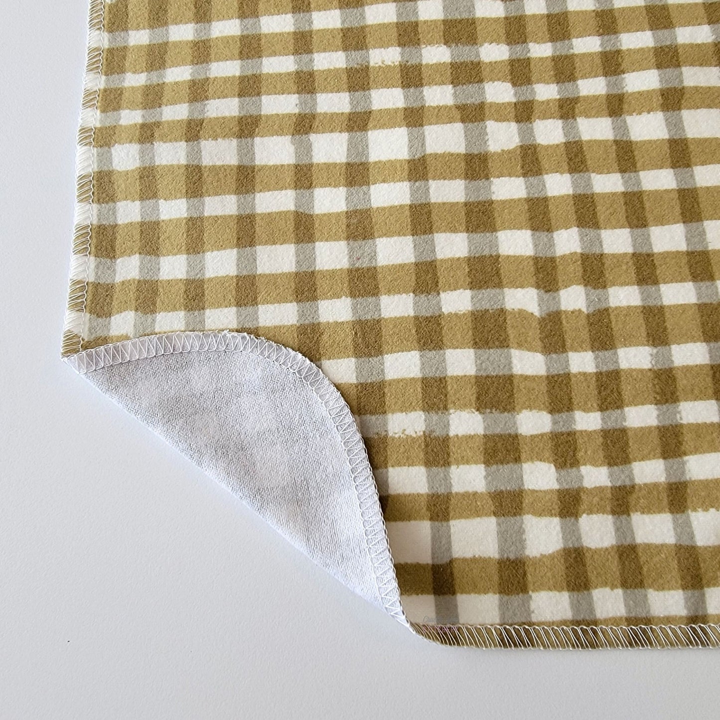 The Olive Painted Plaid NonPaper Towel, shown close-up with the bottom corner folded back to show they are unprinted on the back side, and are one layer.