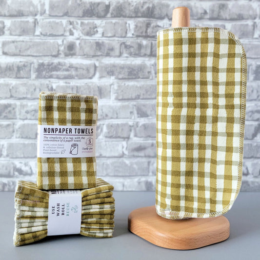 Olive Painted Plaid NonPaper Towels, displayed on a wooden paper towel holder next to packaged bundles. The print is an olive green and cream plaid pattern, and the lines are abstract as if painted.