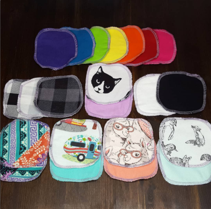 Various reusable cotton rounds arranged on a dark wood table. Rainbow colors, plaid, black cat, bold geometric print, a camper, llamas, animals, and other solid colors.