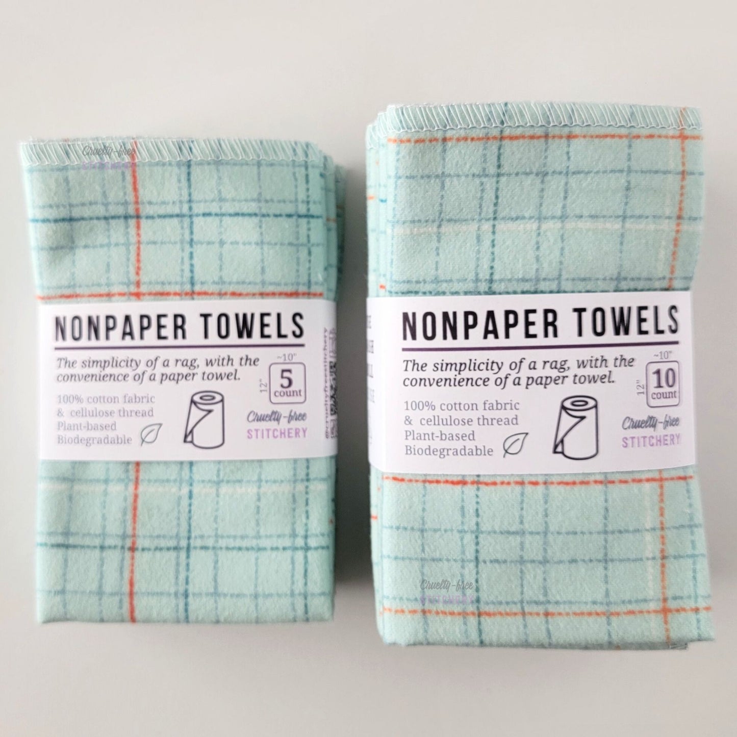 Two packages of the Pale Blue Grid NonPaper Towels, one 5-pack and one 10-pack. Both wrapped with a paper tag, The tag also has the fiber content, a graphic of a paper towel roll, the Cruelty-Free Stitchery logo, and a rectangle with rounded corners that has the dimensions on the outside, 10x12", and the number of towels inside.