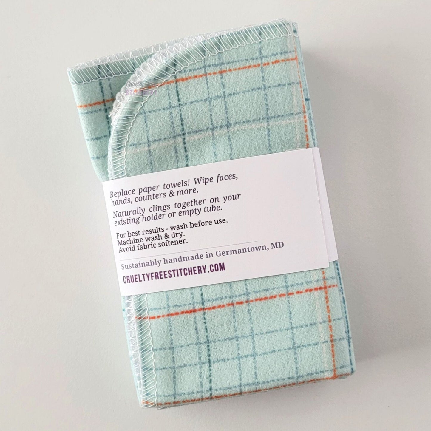 The back of a packaged bundle of the Pale Blue Grid NonPaper Towels. The tag reads "Replace paper towels! Wipe faces, hands, counters, and more. Naturally clings together on your existing holder or empty tube. For best results - wash before use. Machine wash and dry. Avoid fabric softener. Sustainable handmade in Germantown, Maryland. CrueltyFreeStitchery dot com."
