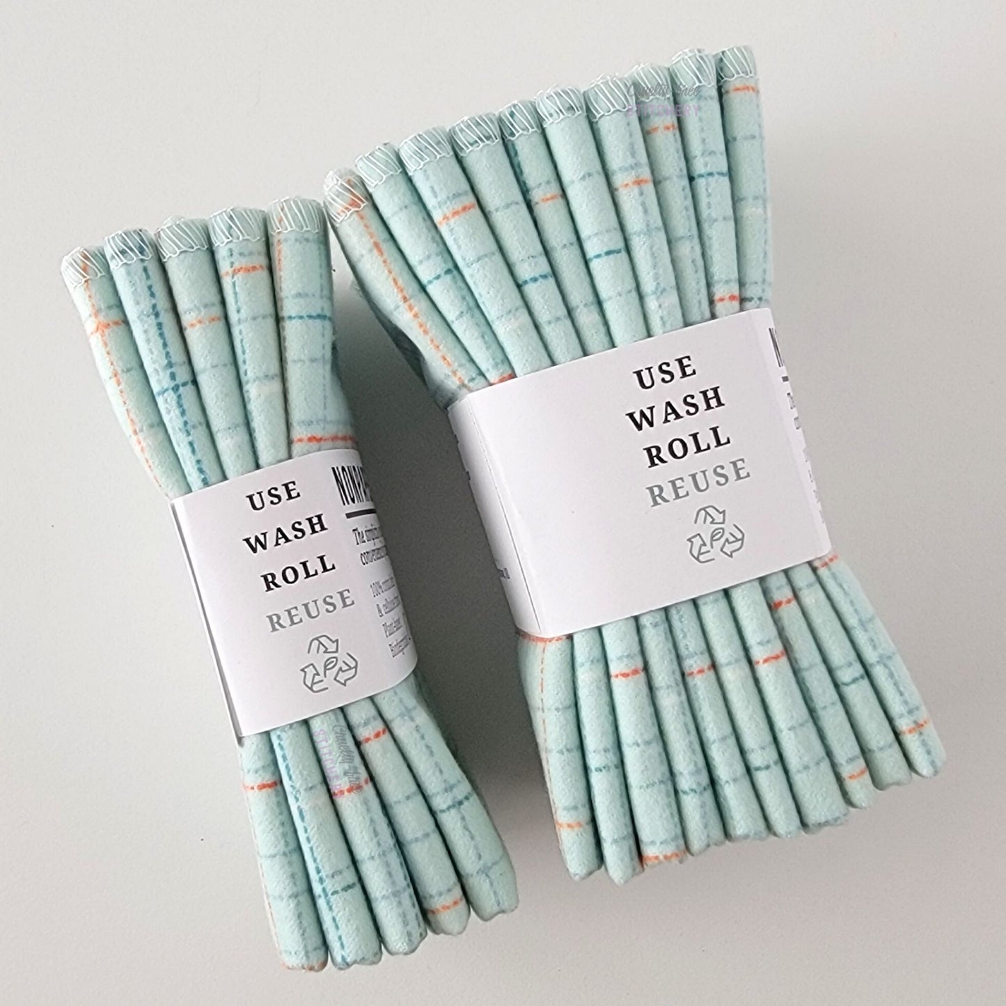 Two packages of the Pale Blue Grid NonPaper Towels, shown on their side to display the 5-pack and 10-packs. The side of the paper tag says "use, wash, roll, reuse" and has the recycling logo with a leaf in the center, to symbolize they are biodegradable.