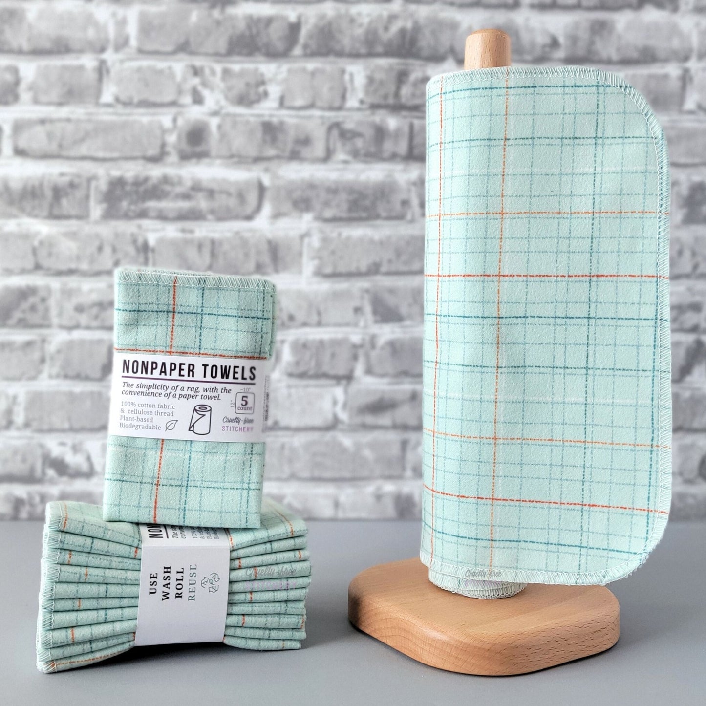 Pale Blue Grid NonPaper Towels, displayed on a wooden paper towel holder next to packaged bundles. The print is a very light blue that almost looks green, with a dotted and dashed irregular grid pattern with thin lines in red, orange, white, darker blue, and gray. 