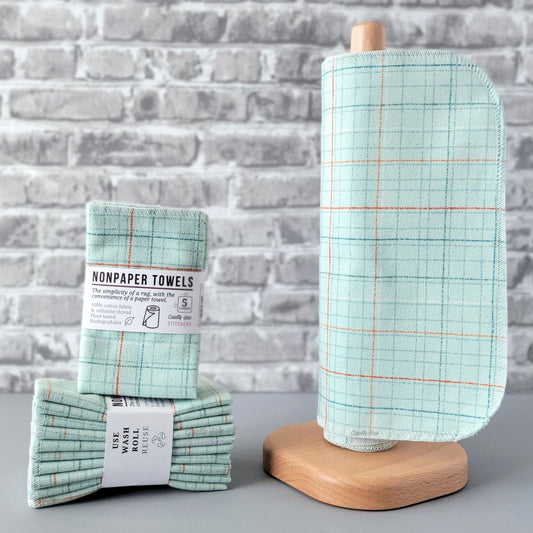 Pale Blue Grid NonPaper Towels, displayed on a wooden paper towel holder next to packaged bundles. The print is a very light blue that almost looks green, with a dotted and dashed irregular grid pattern with thin lines in red, orange, white, darker blue, and gray. 