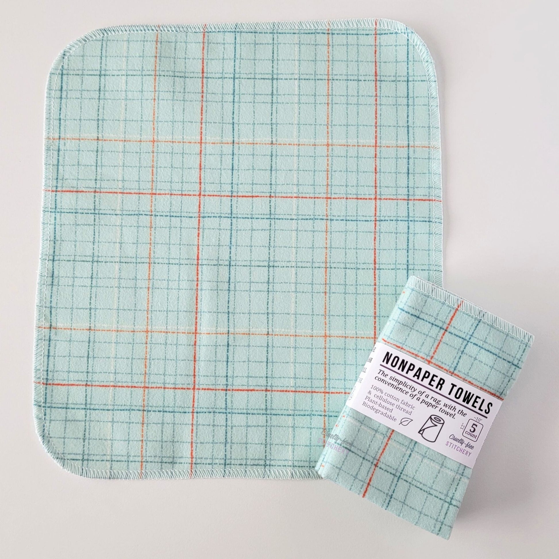 An unfolded Pale Blue Grid NonPaper Towel, next to a bundled 5-pack. The towel is a rectangle with rounded corners, stitched around the edges with white thread. The bundled pack is folded up with a paper wrapper that says "NonPaper Towels, the simplicity of a rag with the convenience of a paper towel"