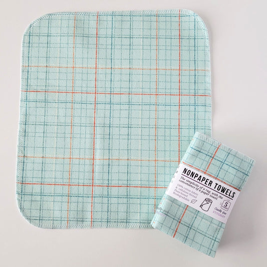 An unfolded Pale Blue Grid NonPaper Towel, next to a bundled 5-pack. The towel is a rectangle with rounded corners, stitched around the edges with white thread. The bundled pack is folded up with a paper wrapper that says "NonPaper Towels, the simplicity of a rag with the convenience of a paper towel"