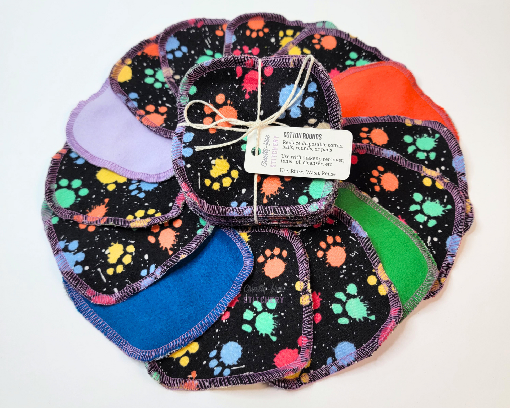Paw print paint splatter cotton rounds. A bundled pack of them on top of another pack arranged in a circle. The front side is black with multicolor splatter paw prints, and the backs are random solid colors, pictured are purple, blue, green, orange.