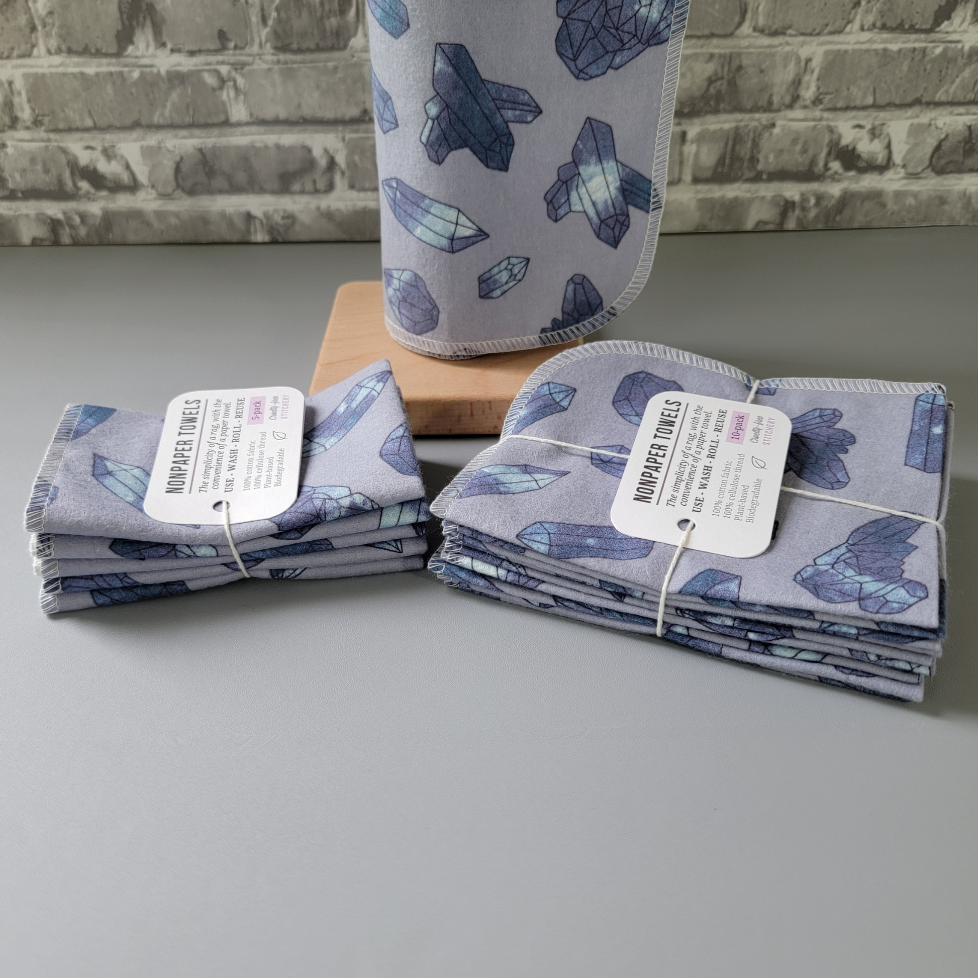 A 5-pack and 10-pack laid next to each other, photographed from the side to show the difference. The 10-pack has the towels folded in half twice, while the 5-pack is folded one more time, making them look smaller. Both are tied with cotton string and a white paper tag.