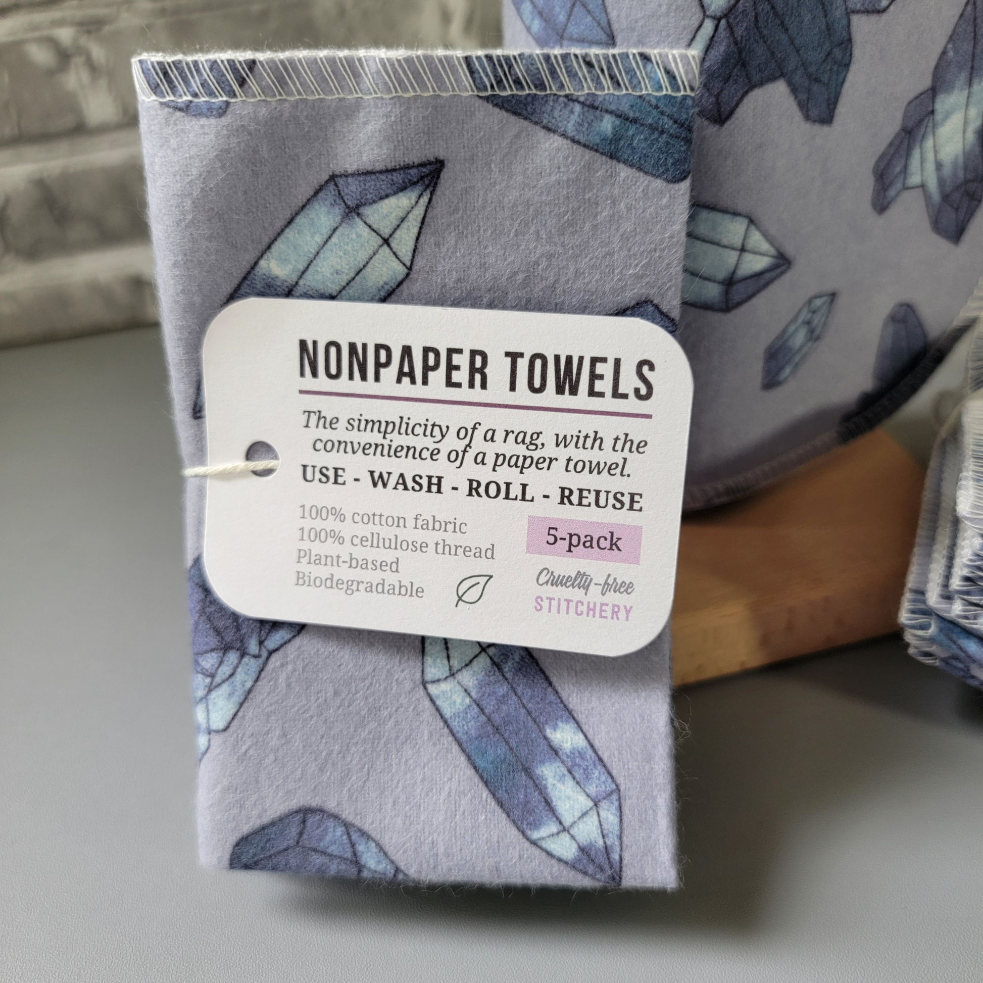 A close-up of the 5-pack, the tag reads "nonpaper towels, the simplicity of a rag with the convenience of a paper towel. Use wash roll reuse. 100% cotton fabric, 100% cellulose thread, plant-based, biodegradable. 5-pack, cruelty-free stitchery" with a tiny green leaf.