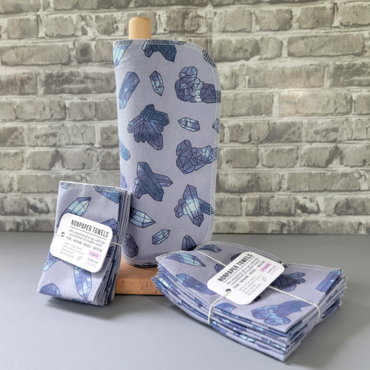 Purple crystal nonpaper towels displayed on a wooden paper towel holder, next to bundled 5 and 10-packs. The towels are a light lavender purple with various sized crystals printed on. The crystals are a tie dye or space effect with black outline. 