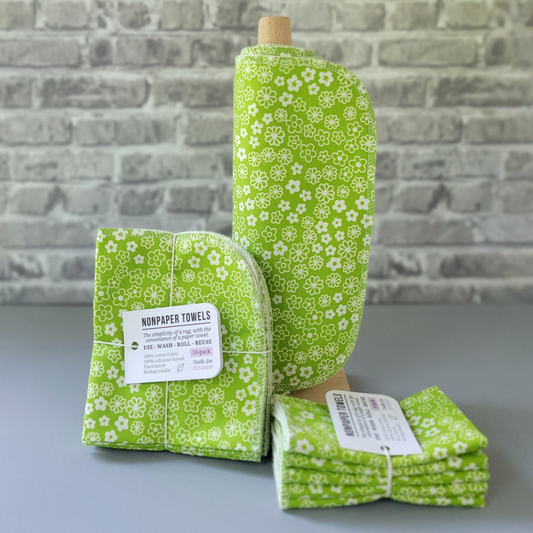 Retro Green Daisy print NonPaper Towels. The print is tiny white simple flowers, some filled on and some outlines, on a bright lime green background. In the center, they are rolled onto a wooden paper towel holder, and there are bundled packs of 10 and 5 sitting next to the holder. The bundles are tied with string and have a small paper tag. The background is a gray brick wall and solid gray tabletop.