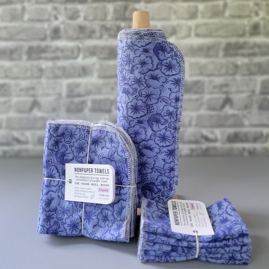Violets print NonPaper Towels. The print is monochrome violet blue, with closely-packed outlined violet flowers. In the center, they are rolled onto a wooden paper towel holder, and there are bundled packs of 10 and 5 sitting next to the holder. The bundles are tied with string and have a small paper tag. The background is a gray brick wall and solid gray tabletop. 