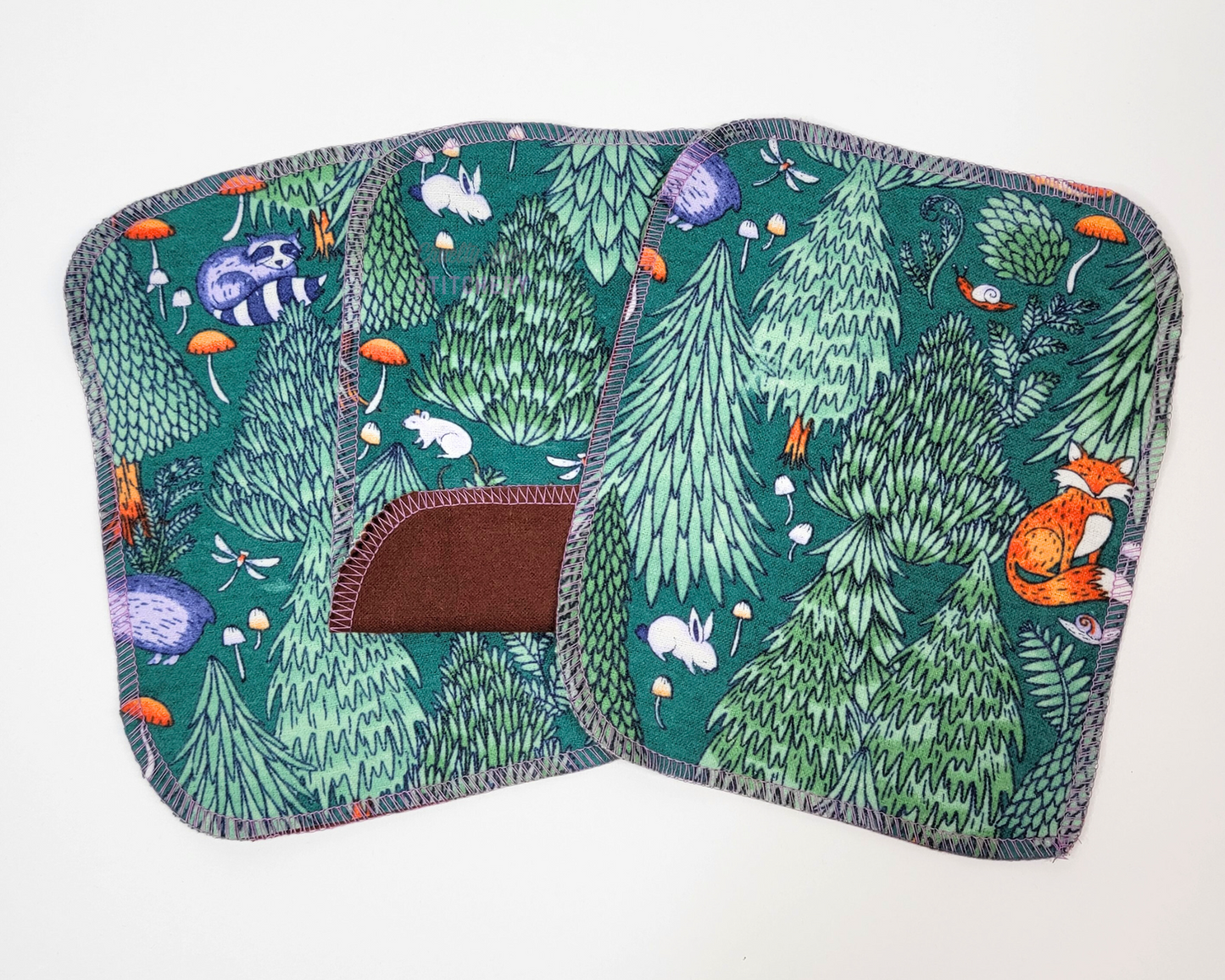 Three reusable cloth wipes in the woodland print. They are a dark green with printed trees, mushrooms, and animals like foxes, raccoons, rabbits, etc. The one in the center is folded up to show that the back side is dark brown.