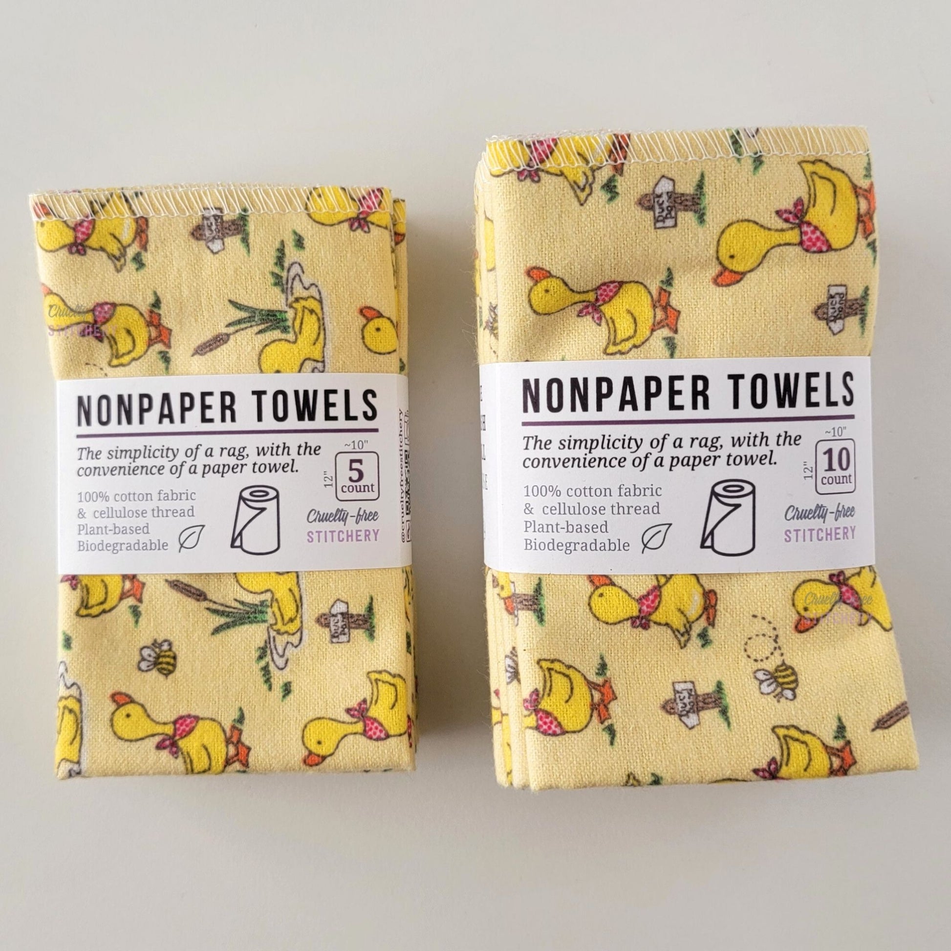 Two packages of the Yellow Ducklings NonPaper Towels, one 5-pack and one 10-pack. Both wrapped with a paper tag, The tag also has the fiber content, a graphic of a paper towel roll, the Cruelty-Free Stitchery logo, and a rectangle with rounded corners that has the dimensions on the outside, 10x12", and the number of towels inside.