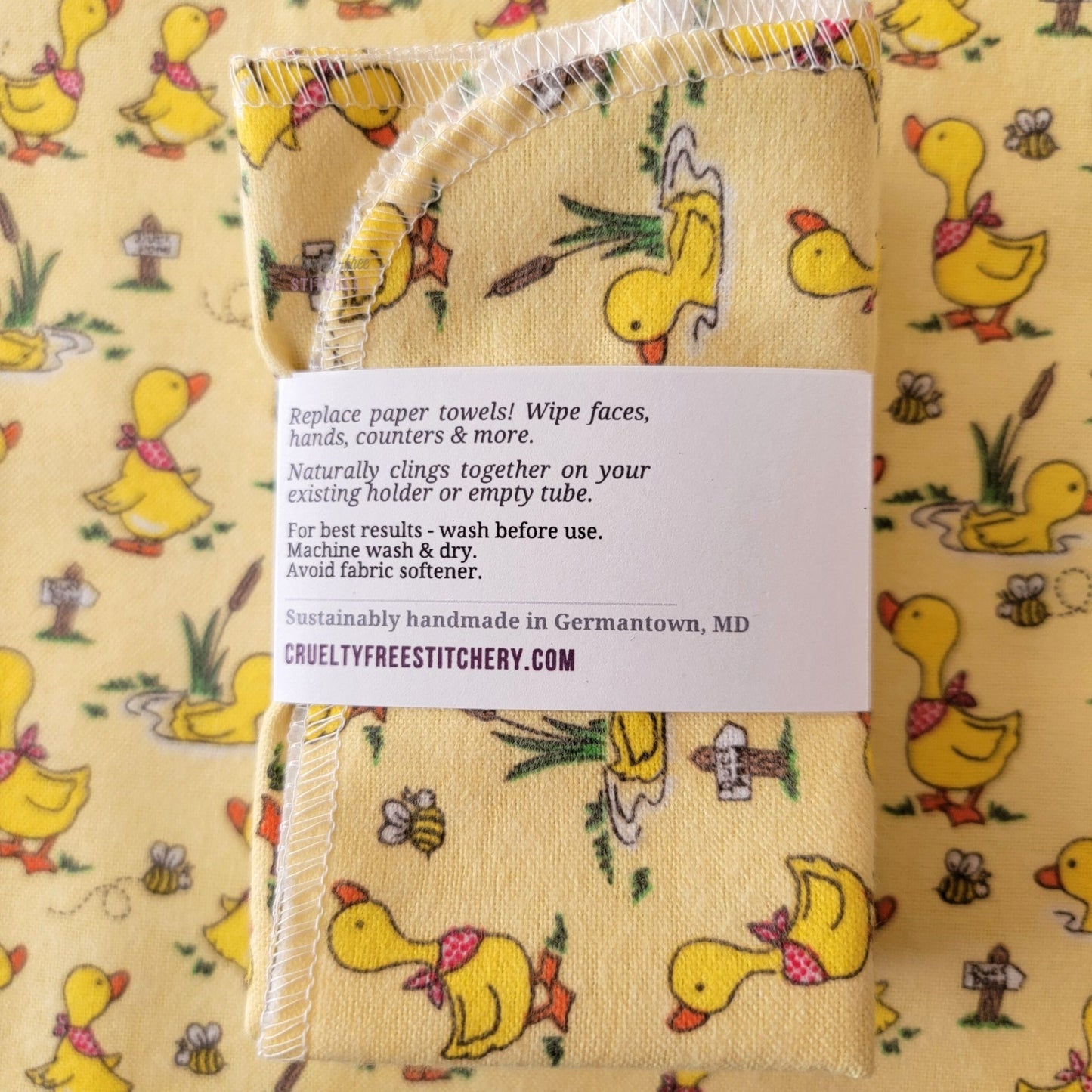 The back of a packaged bundle of the Yellow Ducklings NonPaper Towels. The tag reads "Replace paper towels! Wipe faces, hands, counters, and more. Naturally clings together on your existing holder or empty tube. For best results - wash before use. Machine wash and dry. Avoid fabric softener. Sustainable handmade in Germantown, Maryland. CrueltyFreeStitchery dot com."