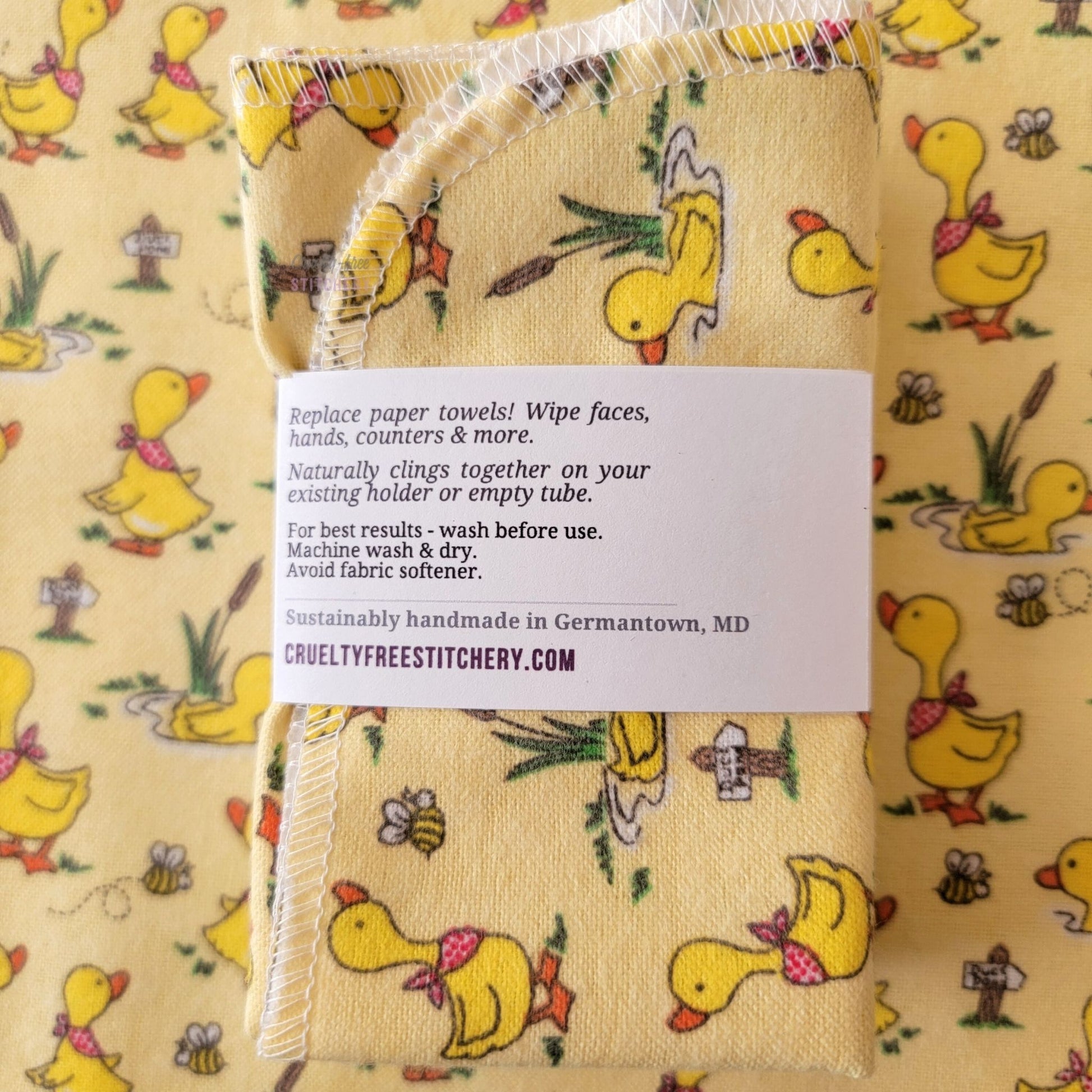 The back of a packaged bundle of the Yellow Ducklings NonPaper Towels. The tag reads "Replace paper towels! Wipe faces, hands, counters, and more. Naturally clings together on your existing holder or empty tube. For best results - wash before use. Machine wash and dry. Avoid fabric softener. Sustainable handmade in Germantown, Maryland. CrueltyFreeStitchery dot com."