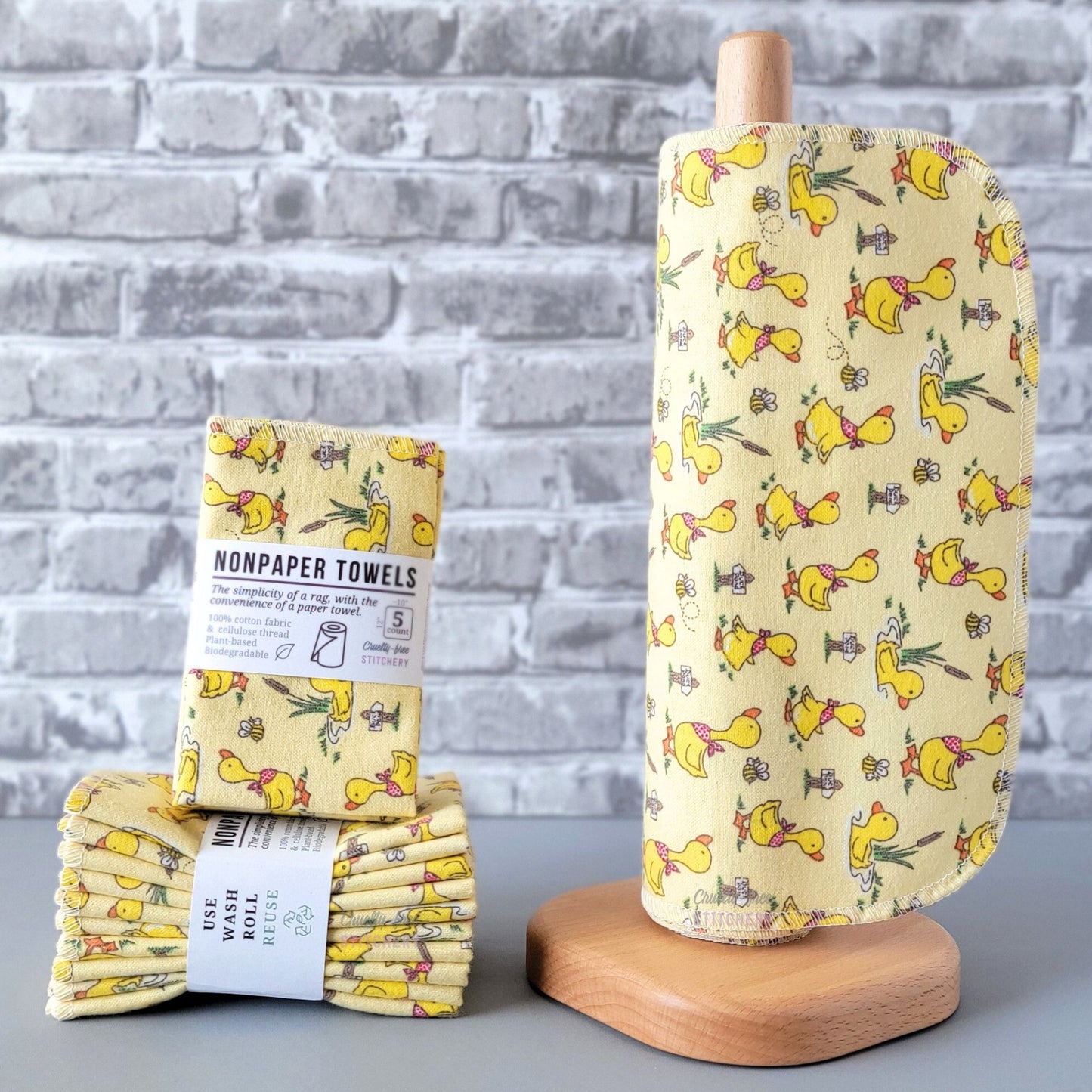 Yellow Ducklings and Bees NonPaper Towels, displayed on a wooden paper towel holder next to packaged bundles. The print is a pale yellow background with bright yellow ducklings wearing red spotted bandanas, walking around and swimming. There are cattails, tiny bumblebees, and little wooden signs that say "duck pond" scattered around as well. The ducks are a little over an inch tall. 