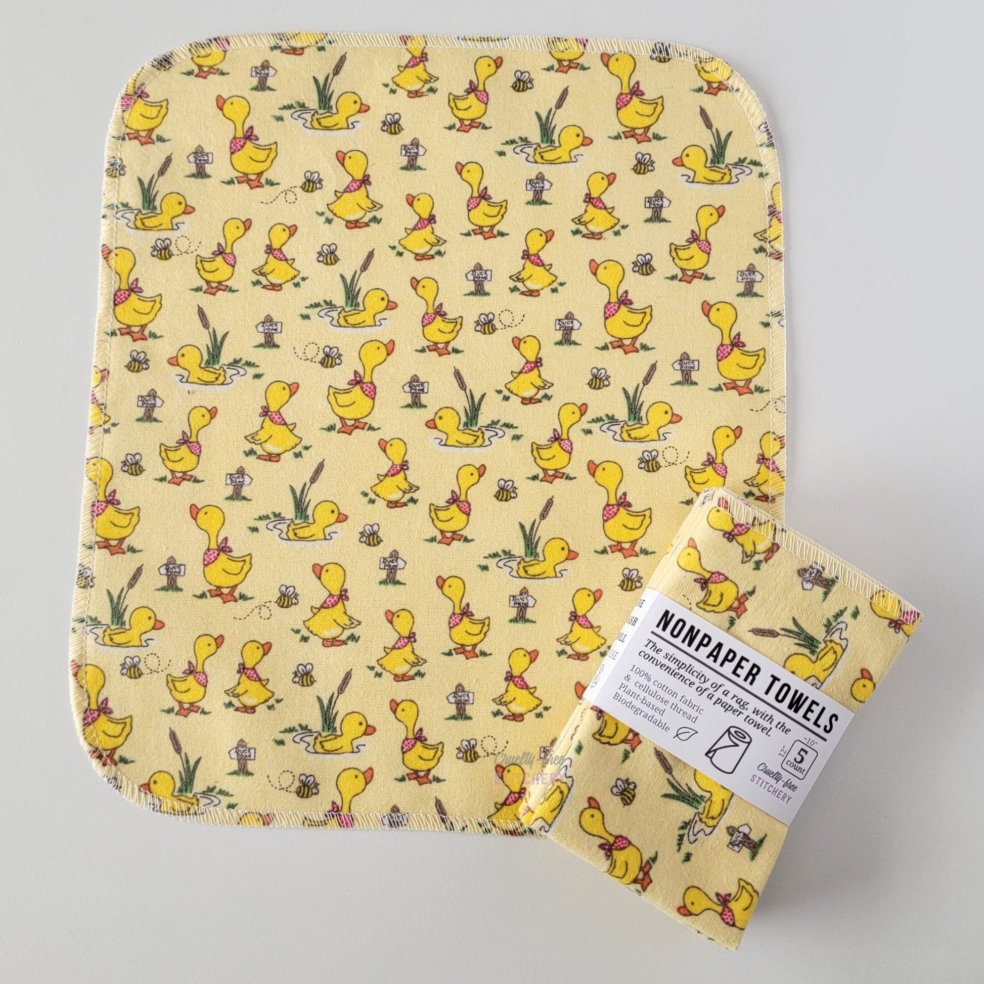 An unfolded Yellow Ducklings NonPaper Towel, next to a bundled 5-pack. The towel is a rectangle with rounded corners, stitched around the edges with white thread. The bundled pack is folded up with a paper wrapper that says "NonPaper Towels, the simplicity of a rag with the convenience of a paper towel"

