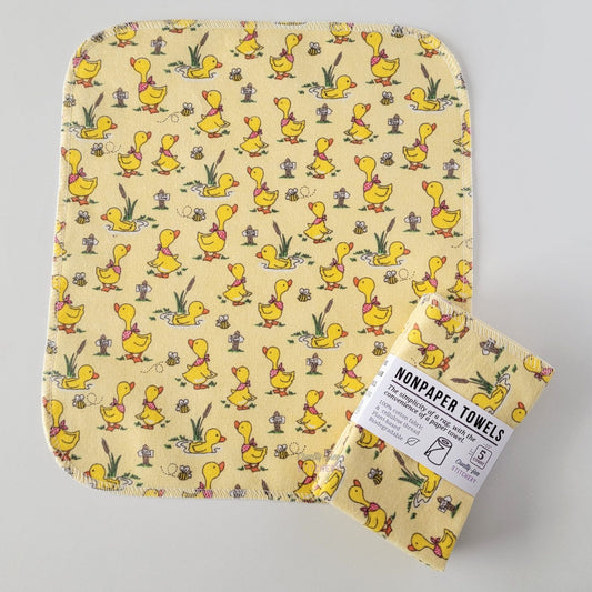 An unfolded Yellow Ducklings NonPaper Towel, next to a bundled 5-pack. The towel is a rectangle with rounded corners, stitched around the edges with white thread. The bundled pack is folded up with a paper wrapper that says "NonPaper Towels, the simplicity of a rag with the convenience of a paper towel"
