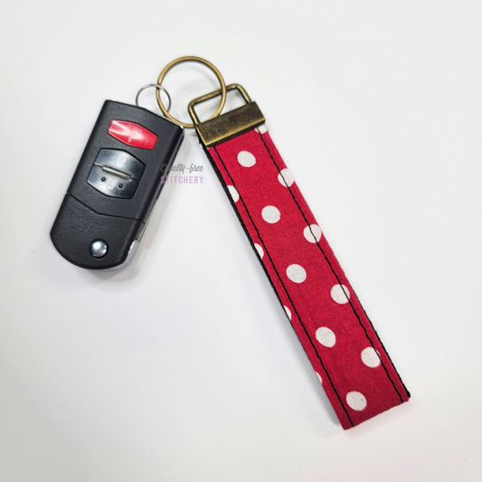 Red with white polka dots print key fob wristlet attached to a car key for scale. The car key is the fold-away type and is about half as long as the strap.