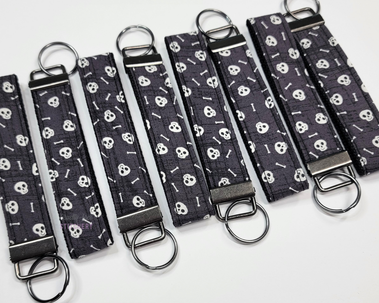 Wristlet key fobs arranged in a row of eight with ends alternating. The wristlet is dark grey with tiny white skulls and bones. The hardware and key ring are gunmetal silver.