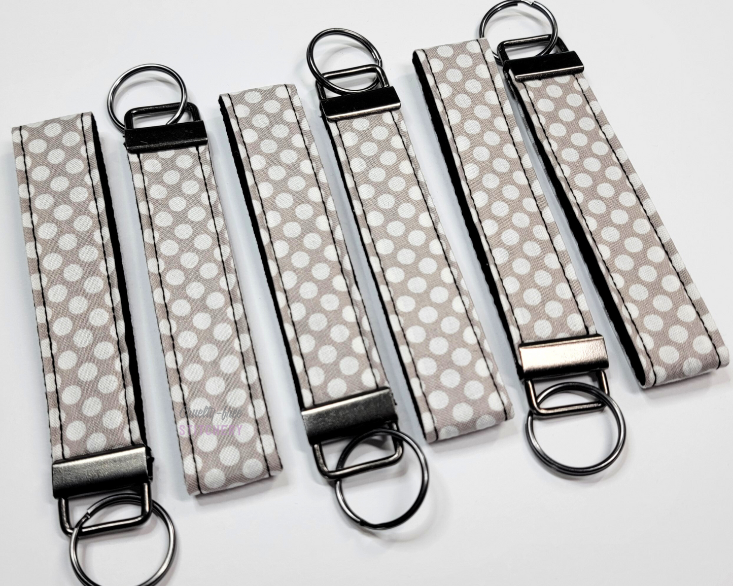 Wristlet key fobs arranged in a row of six with ends alternating. The wristlet is light grey fabric with closely printed white dots. The hardware and key ring are gunmetal silver.