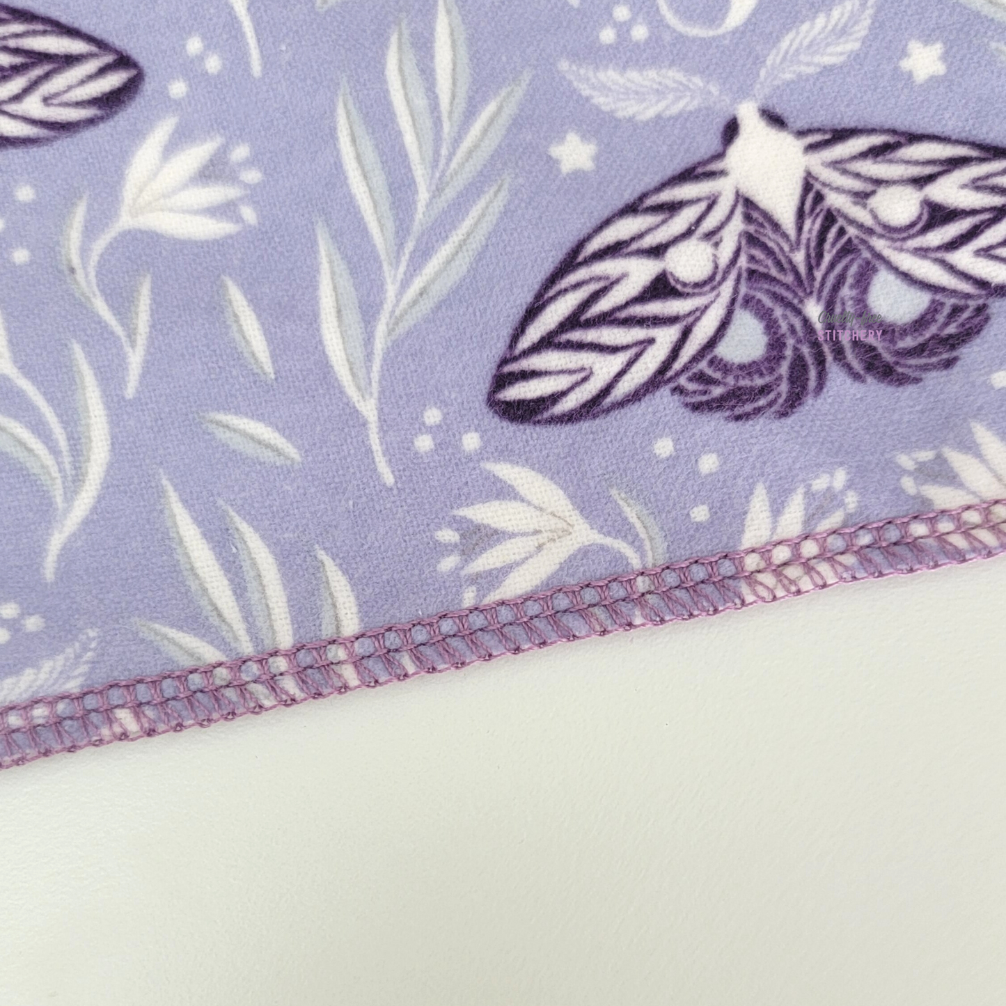 Close-up of the bottom edge of the purple moths NonPaper Towel. The towel has a light purple background with white vines and dark purple moths, and the edge is stitched with a lilac purple thread.