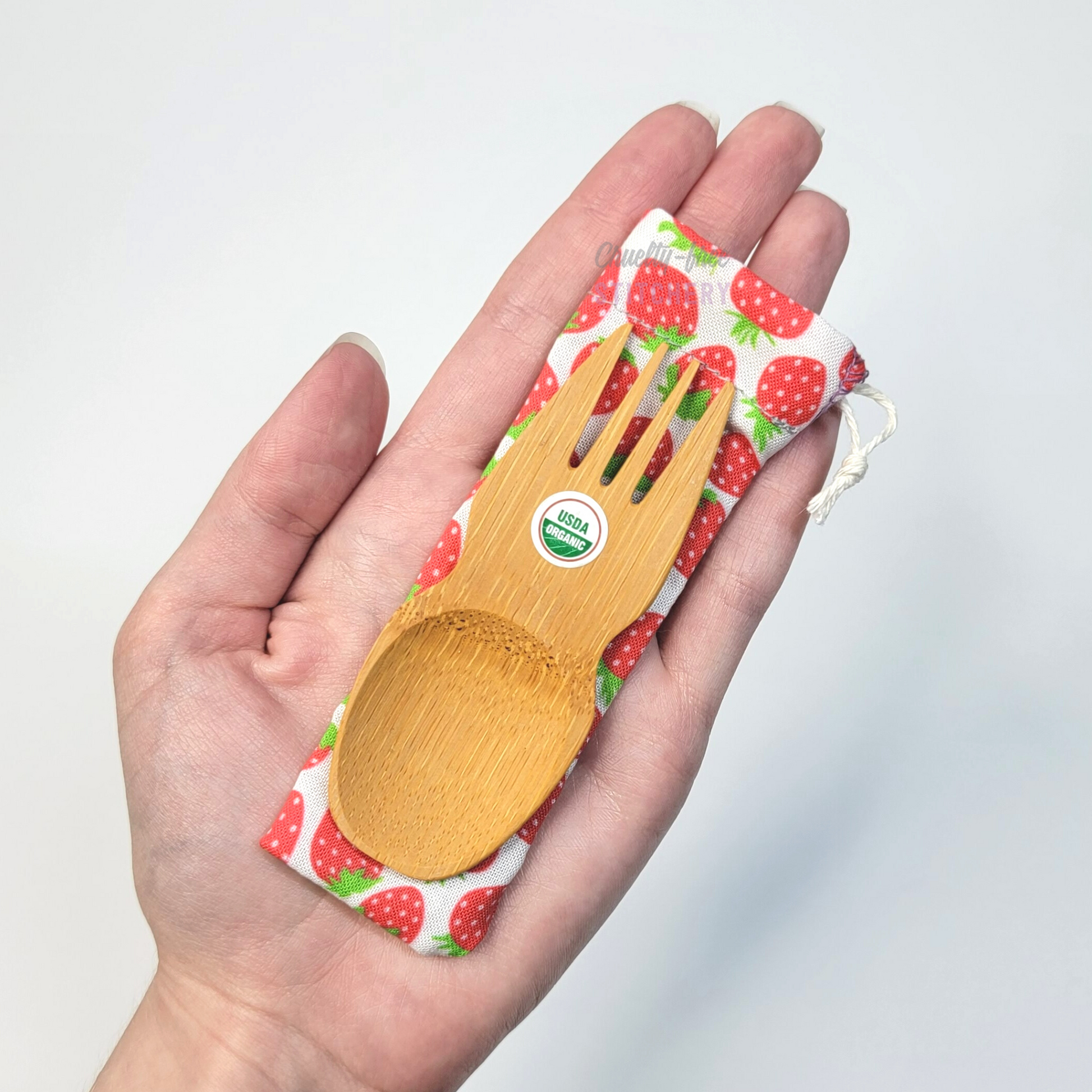 Strawberry print fabric pouch with bamboo spork on top, laid on a hand for size reference. The spork is slightly longer than the fingers.