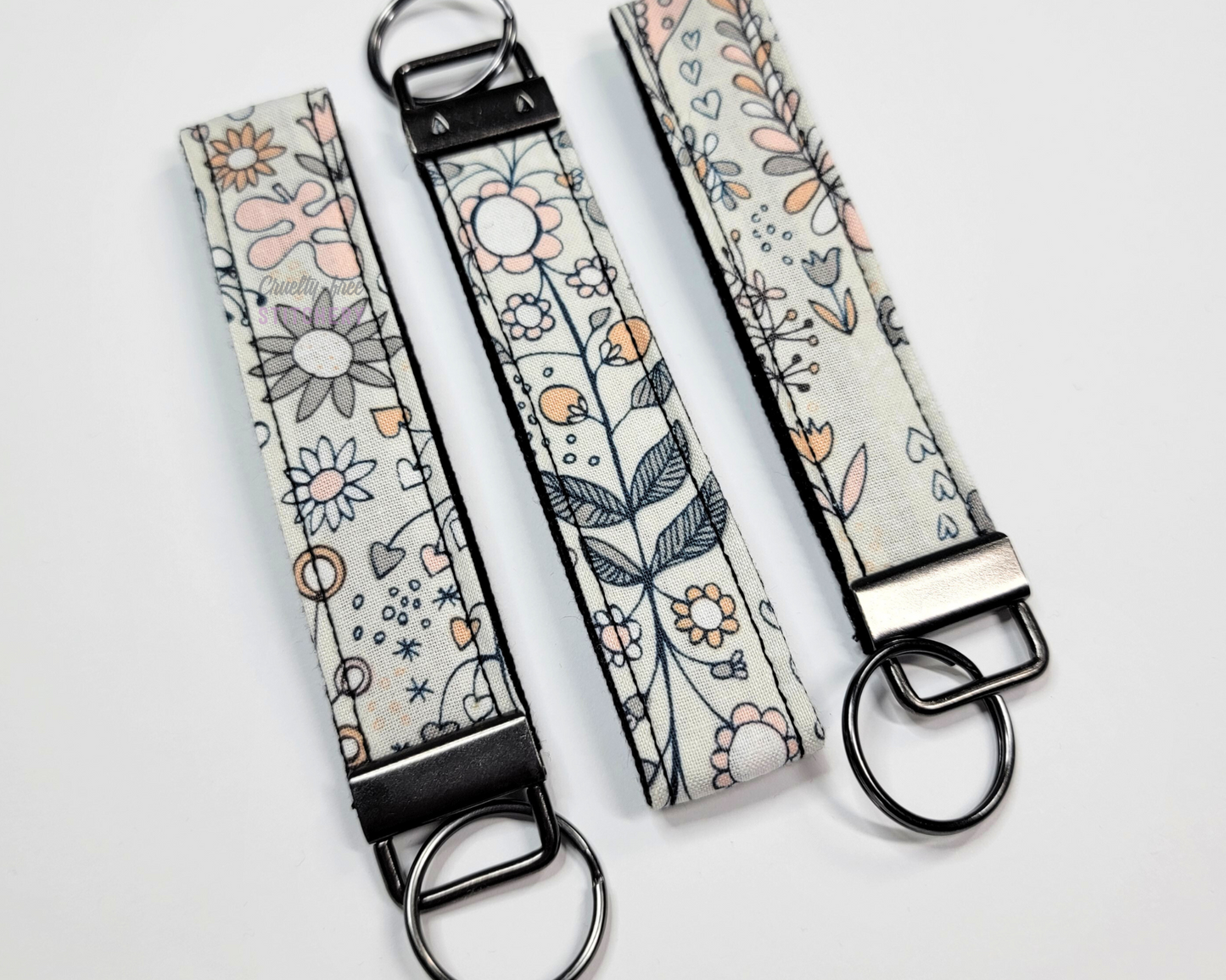 Wristlet key fobs arranged in a row of three with ends alternating. The wristlet is a light grey fabric with whimsical doodled pink and dark grey flowers. The hardware and key ring are gunmetal silver.