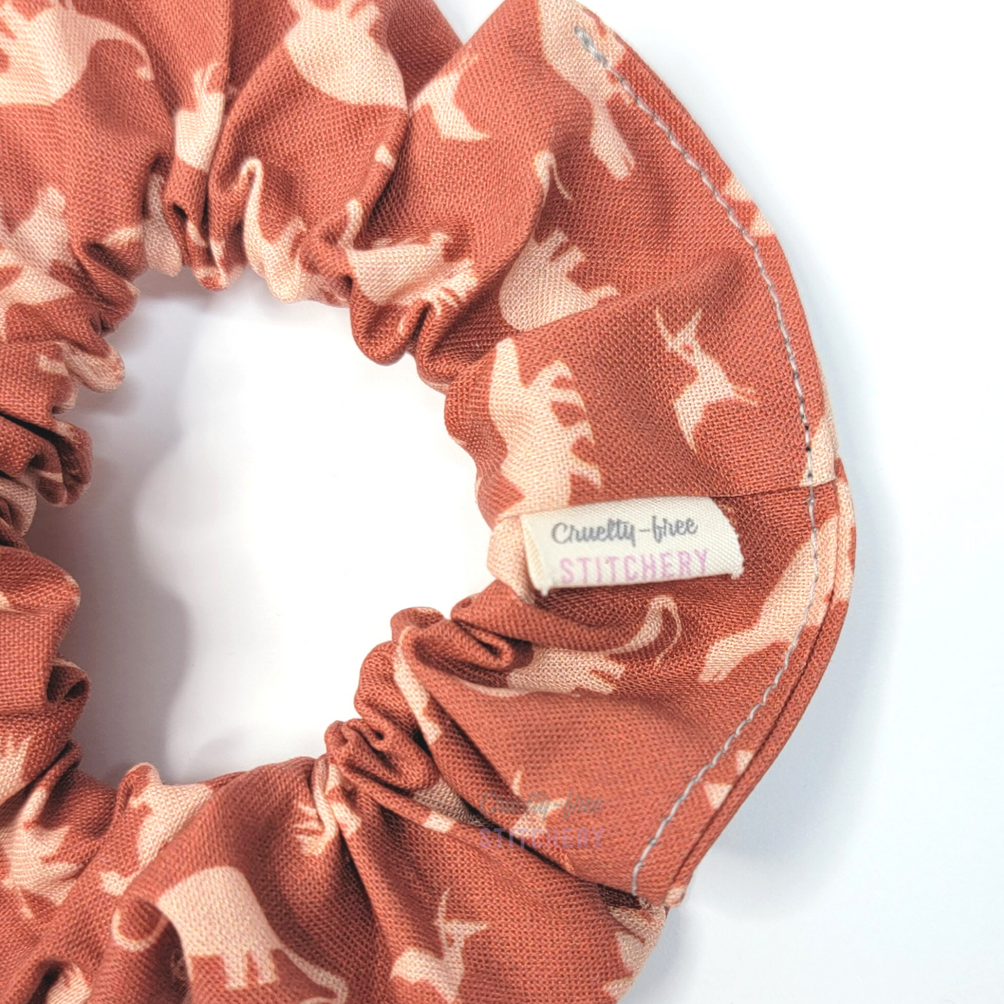 Dark orange with light orange dinosaurs print scrunchie - close-up with a small white tag with the Cruelty-Free Stitchery logo.