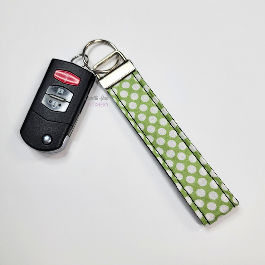 Green with white dots print key fob wristlet attached to a car key for scale. The car key is the fold-away type and is about half as long as the strap.