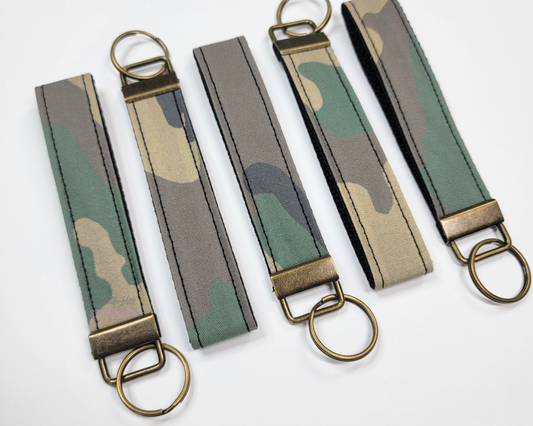 Key fob wristlets lined up in alternating directions. They are a traditional camo print fabric strap with an antiqued brass hardware and key ring. All of the camo patterns are unique.