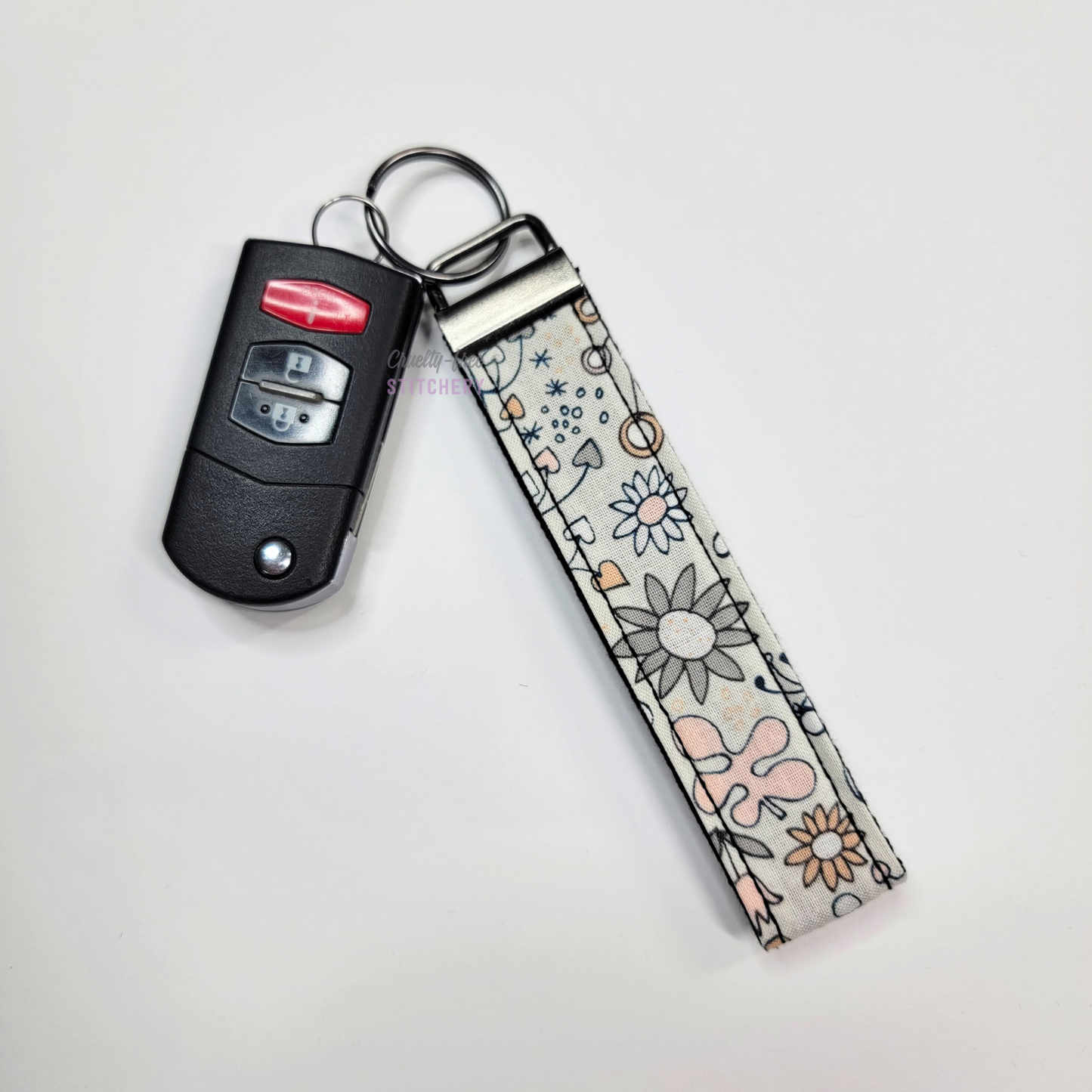 Grey and pink flowers print key fob wristlet attached to a car key for scale. The car key is the fold-away type and is about half as long as the strap.