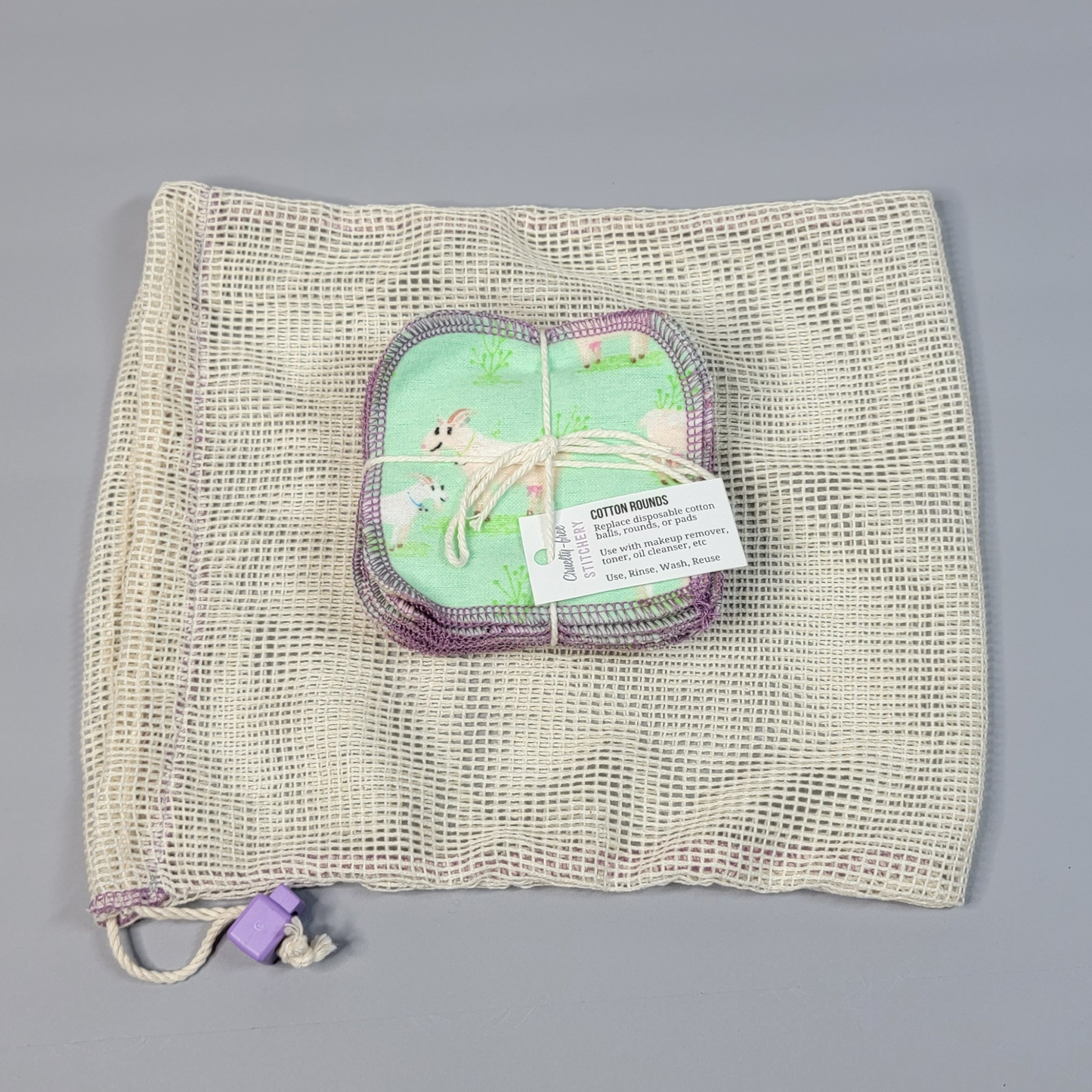 A bundled pack of cotton rounds on top of a mesh drawstring bag. The mesh is a natural off-white color with square holes, and a light purple cord stop.