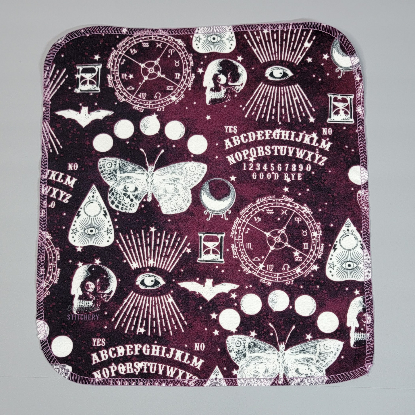 A NonPaper Towel in the Ouija print - a galaxy background with various shades of light and dark burgundy, with white printed motifs like ouija boards, planchettes, moths, skulls, bats, astrological charts, crystal balls, etc.