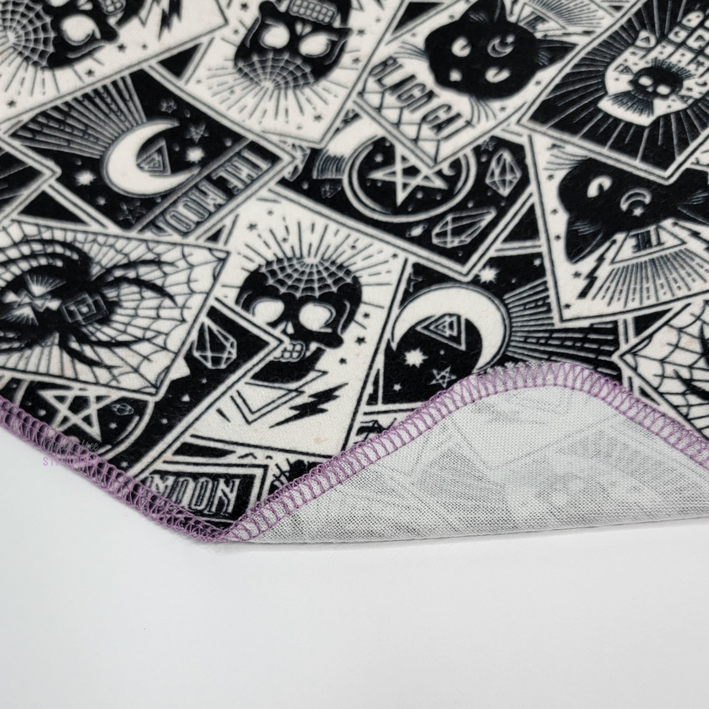 A NonPaper Towel that is white with scattered Tarot cards printed in black, showing various spiders, skulls, and cats. The corner is folded up to show the purple stitching and that the opposite side is white.