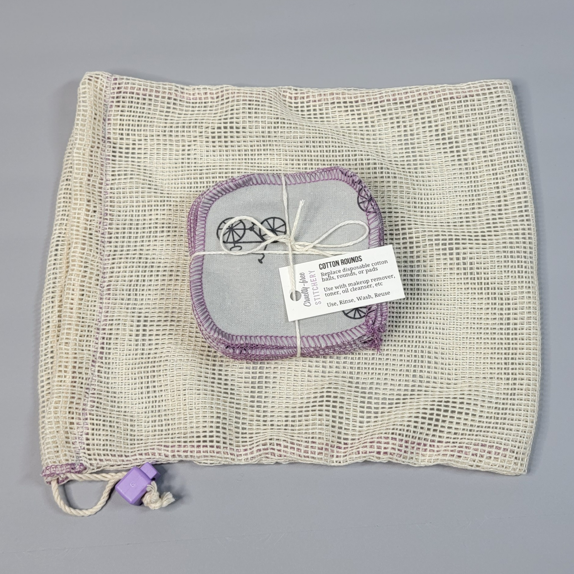 Bundled pack of grey with bicycles cotton rounds on top of a mesh drawstring bag. The bag is a natural off-white colored square mesh, with a light purple cord stop.