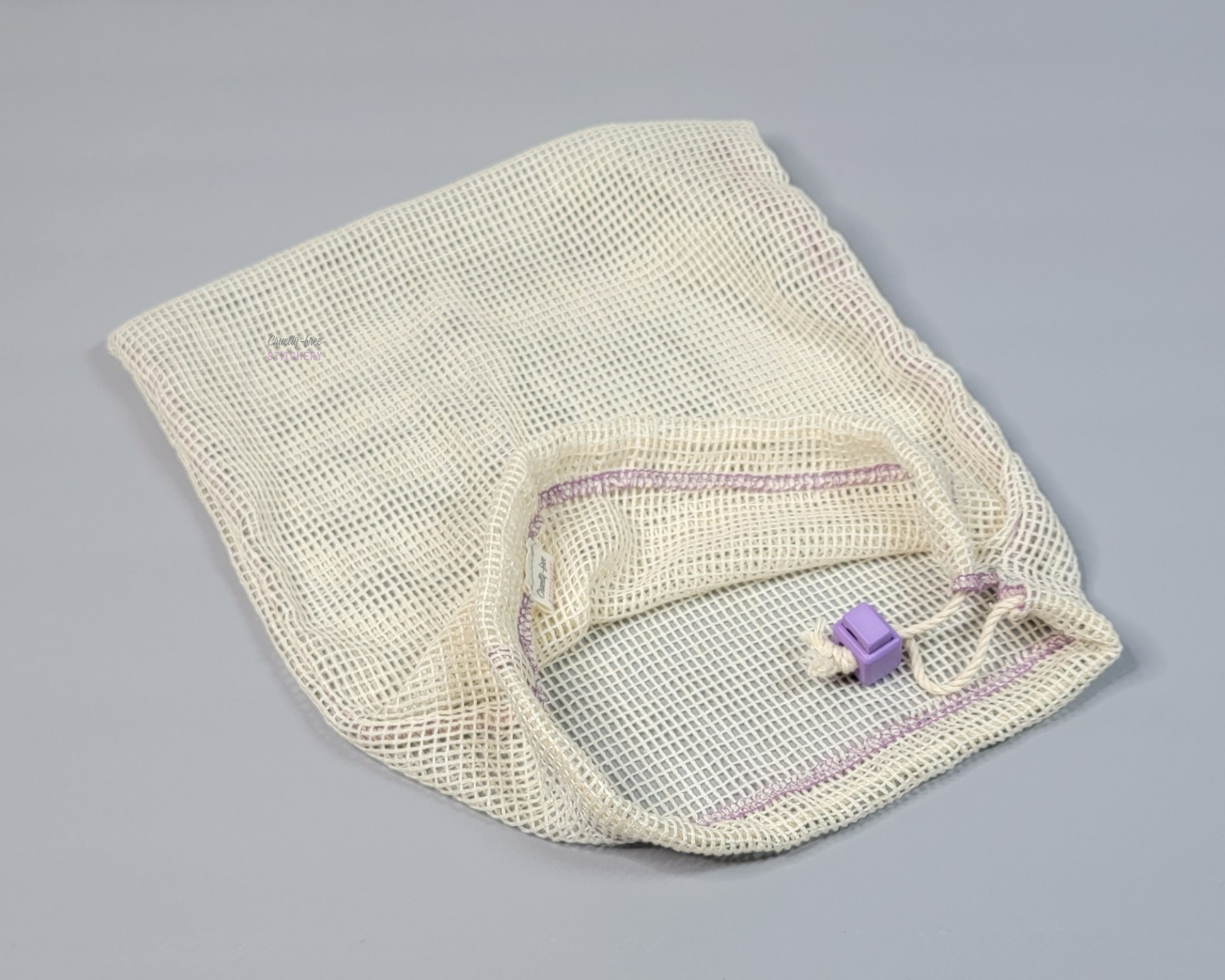 An off-white cotton mesh bag. Drawstring closure with a light purple square cord stop.