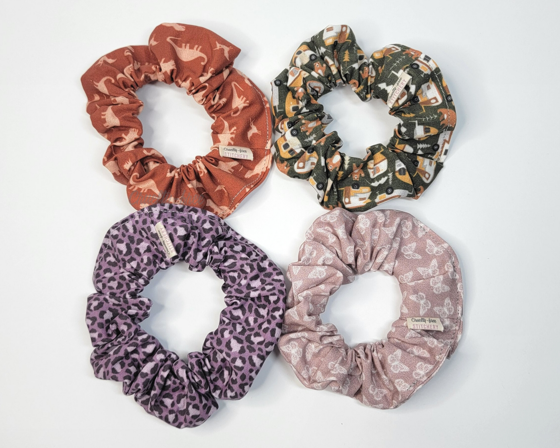 Four scrunchies on a white background. A dark orange with dinosaurs, dark green with campers and bears, violet purple leopard print, and a mauve pink with tiny white butterflies.