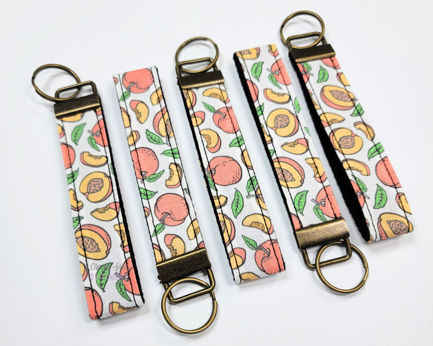 Wristlet key fobs arranged in a row of five with ends alternating. The wristlet is peaches printed on a white background, and the hardware and keyring are an antiqued brass color.