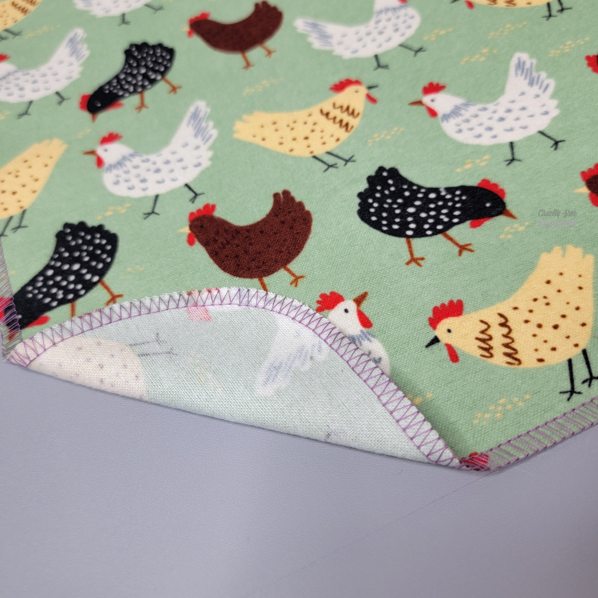 The corner of the green with chickens print NonPaper Towel with the edge folded up to show the back, which is unprinted white. 