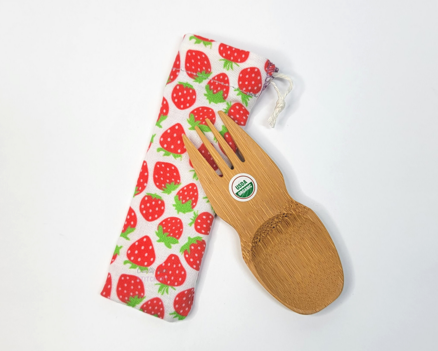 Reusable bamboo spork with pouch. The pouch is a white background with tiny red strawberries. The pouch is sitting diagonally with the spork partially on top pointing the other way. The spork is small, a double ended fork and spoon.