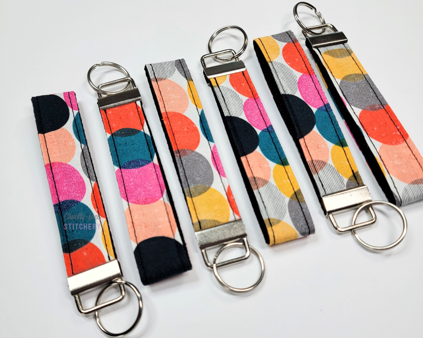 Wristlet key fobs arranged in a row of six with ends alternating. The wristlet is fabric with large multicolored dots - teal, hot pink, yellow, orange, black. The hardware and key ring are silver.