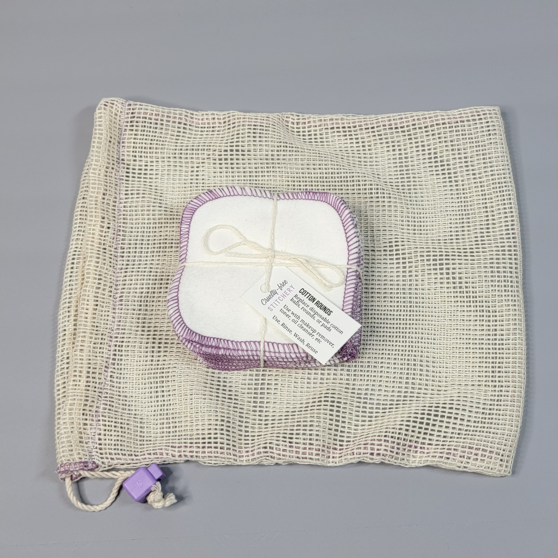 A pack of white reusable cotton rounds on top of a mesh washing bag - the bag is an off-white color with square mesh, and a light purple cord stopper.