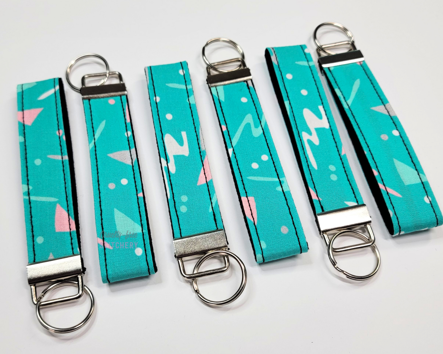 Wristlet key fobs arranged in a row of six with ends alternating. The wristlet is turquoise fabric with pink and grey 90s inspired squiggles. The hardware and keyring are silver.
