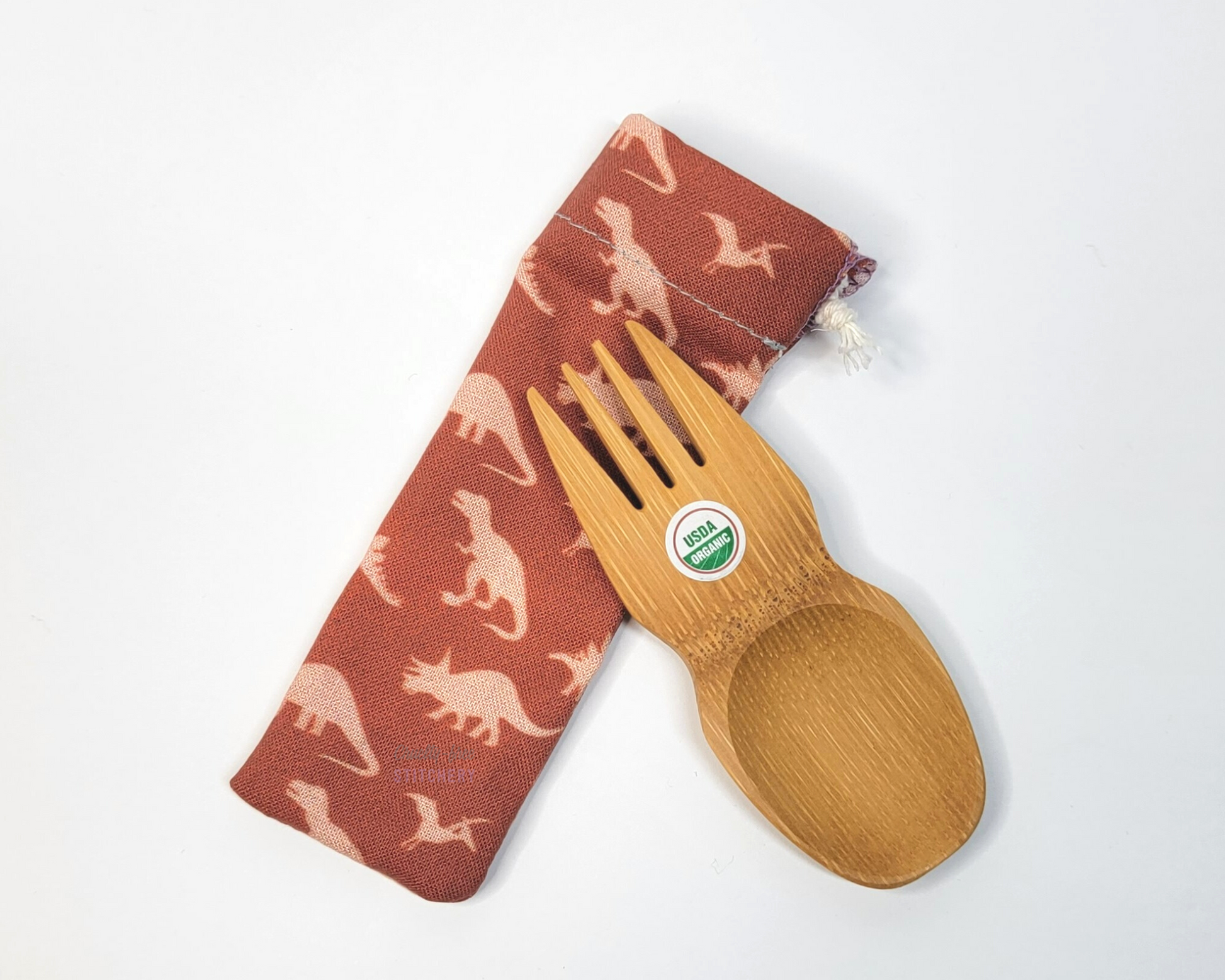 Reusable bamboo spork with pouch. The pouch is a dark orange with light orange dinosaurs. The pouch is sitting diagonally with the spork partially on top pointing the other way. The spork is small, a double ended fork and spoon.