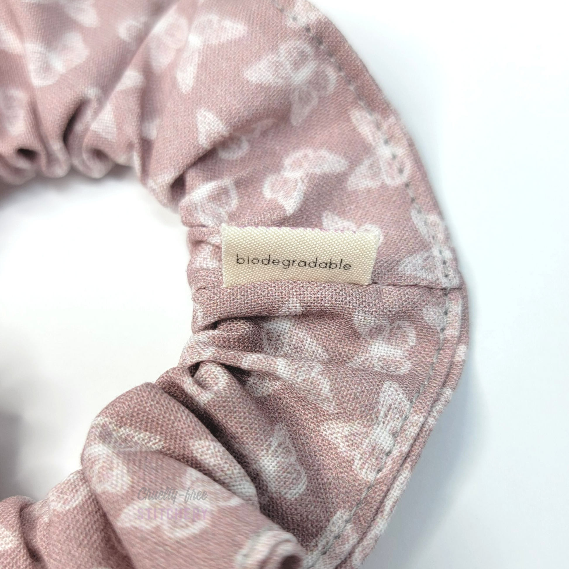 Muted mauve pink scrunchie with tiny white butterflies, The back of the small white tag says "biodegradable"